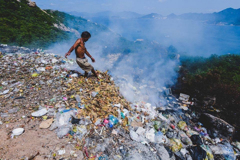 Bức ảnh ghi lại ông Nguyễn Lương, người phụ trách lò đốt rác ở đảo Bình Ba (Khánh Hòa) đang dùng chân đẩy rác. Xung quay cây cối chết khô. Ngay phía dưới là bãi tắm và là vịnh nuôi tôm hùm, thuỷ hải sản.