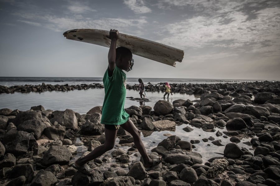 Một cậu bé khiêng ván lướt sóng bị hỏng trên bãi biển tại Pointe des Almadies, ở Dakar - thủ đô đất nước Sénégal. Ảnh: 36 giờ ở Dakar - Finbarr O'Reilly